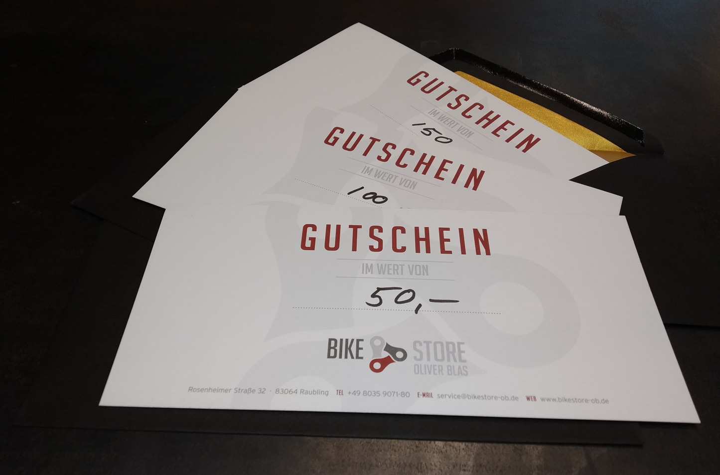 Dreierlei Gutscheine im Wert von 50,-, 100,- und 150,- € zu einer Art Fächer arrangiert auf einem schwarzen Tisch und schwarz/goldenem Kuvert.