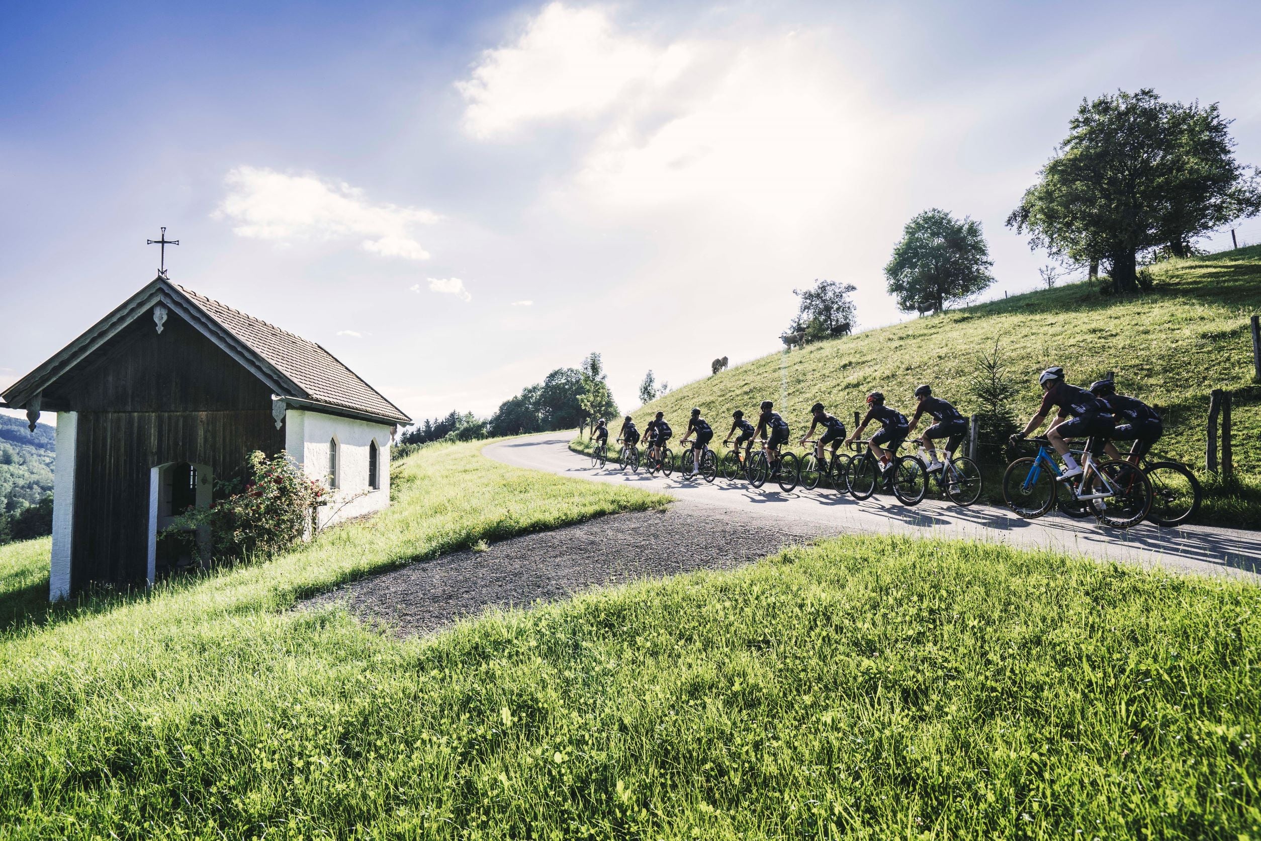 Radfahrer in Kolonne fahren auf einen Berg. Linker Hand eine kleine Kapelle