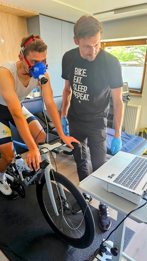 Sportler mit Atemmaske bei einer Leistungsdiagnostik auf einem Rennradtrainer. Daneben steht ein MItarbeiter von Ineofit und überwacht an einem Laptop den Prüflauf.